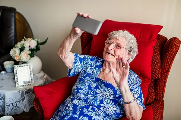 An older woman sits in a red armchair, smiling and holding up a smartphone as if taking a selfie or video call.